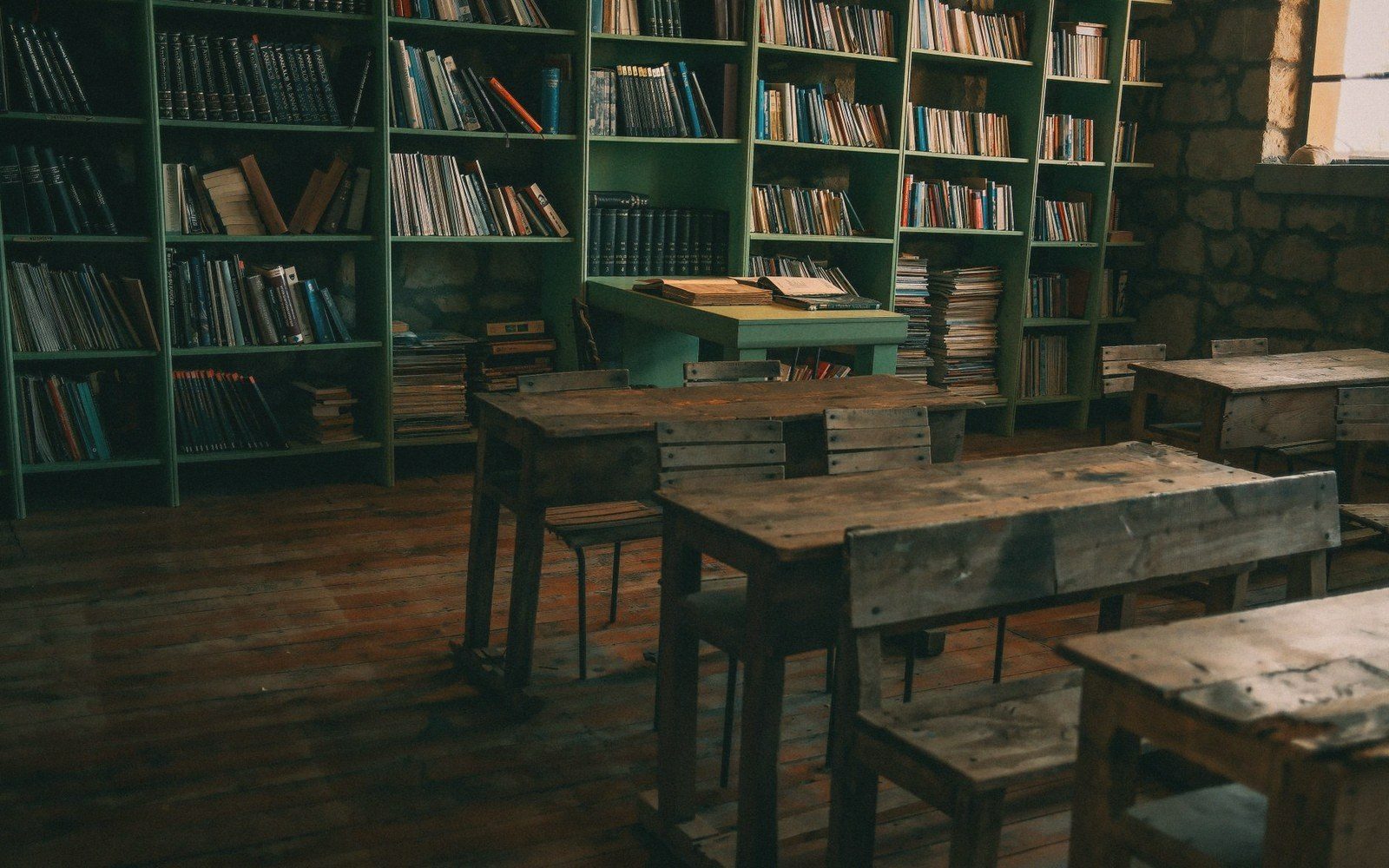 Wooden desks and chairs face green bookshelves packed with books in a dim classroom