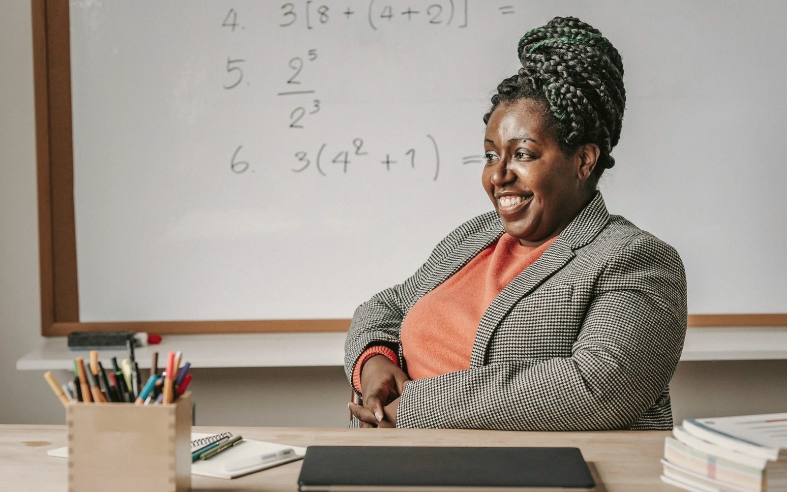 Smiling teacher sits at a desk in front of a whiteboard with math equations