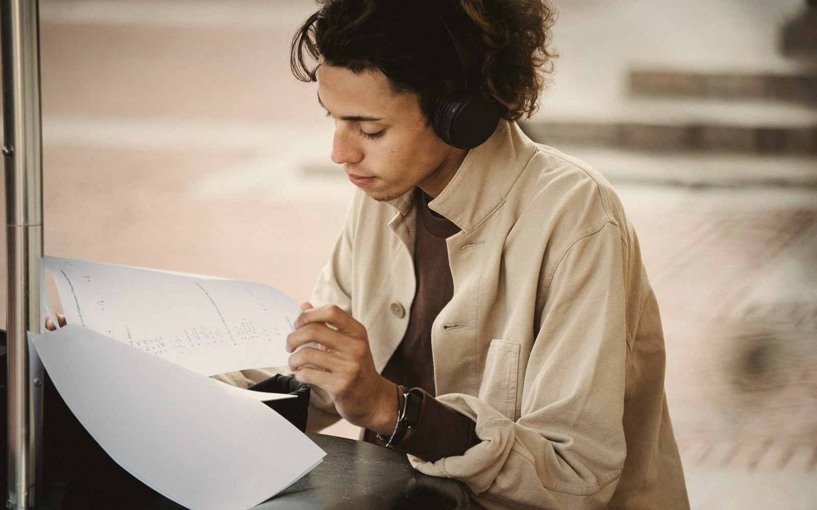 Young man wearing headphones reads sheet music while writing outdoors at a table