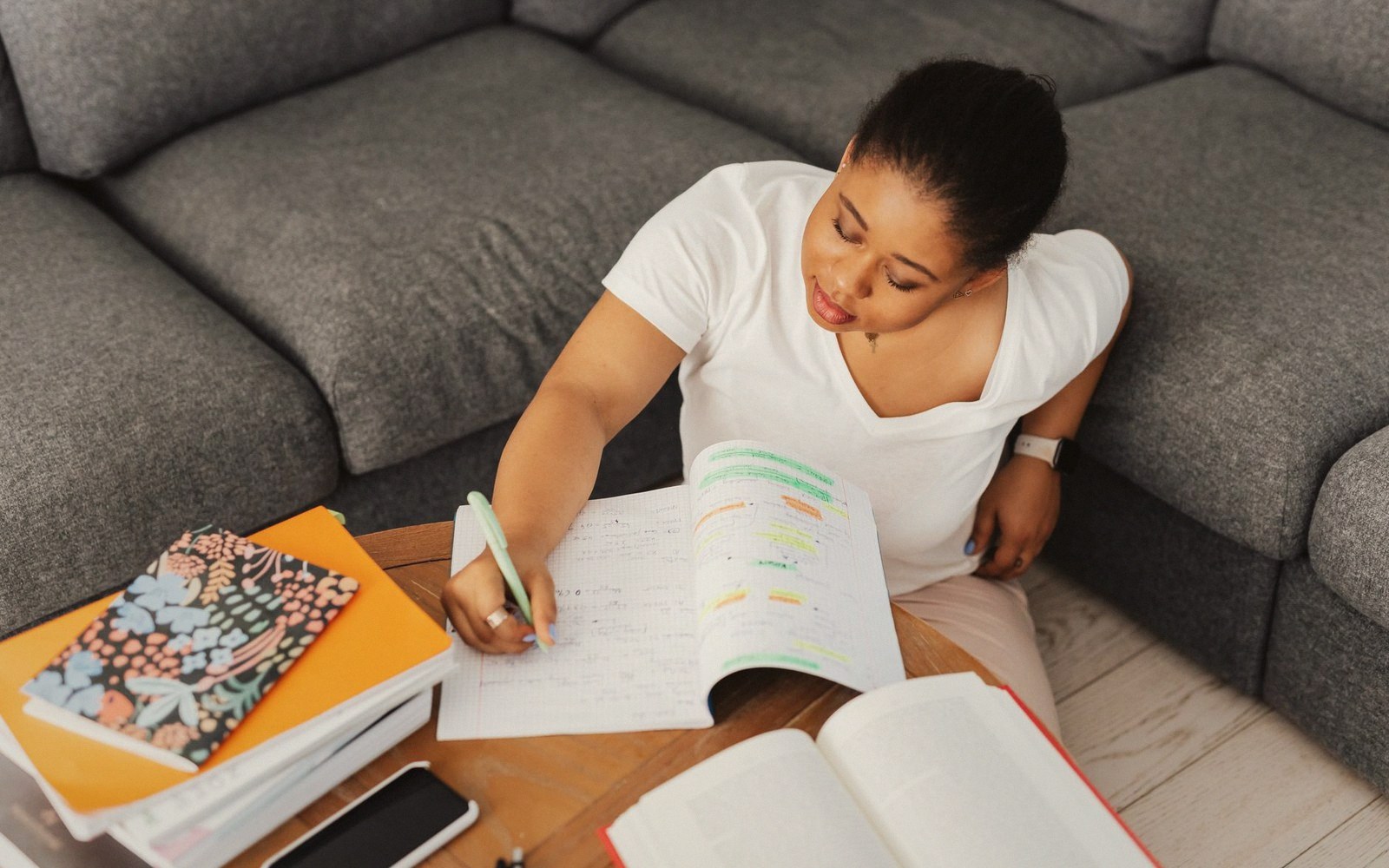 Woman sitting on a couch writing notes in a highlighted textbook on a coffee table