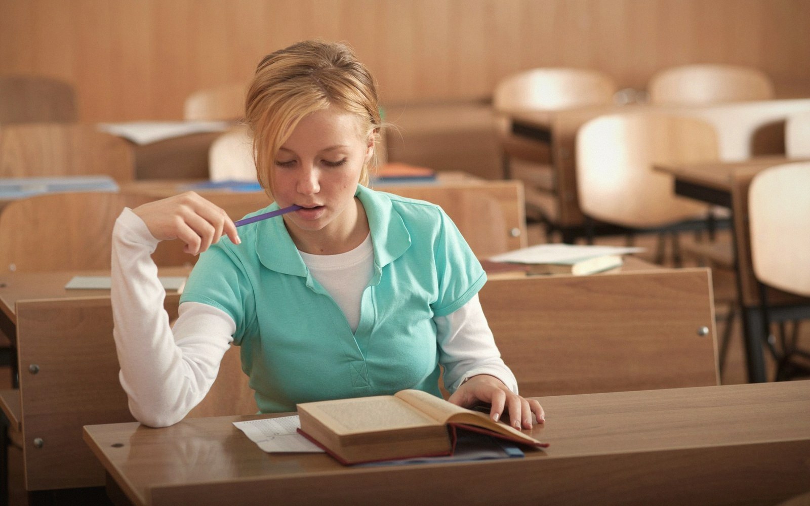 Student chews a pencil while studying a textbook at a classroom desk