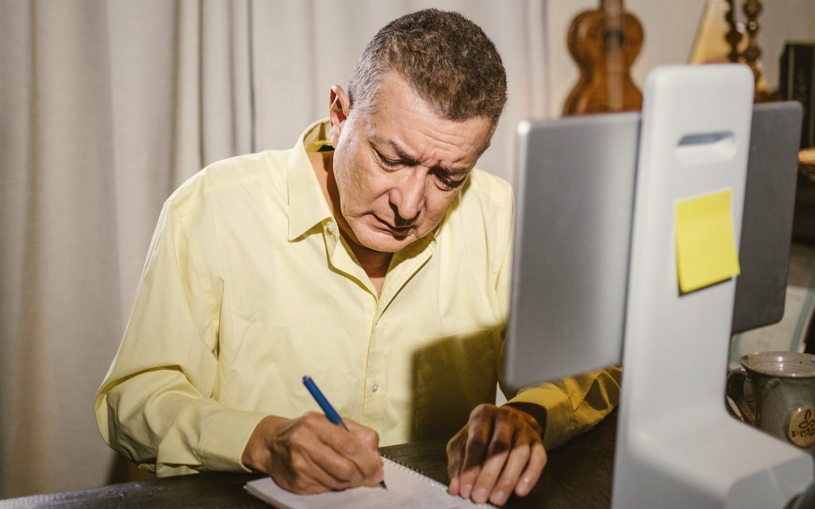 Man in a yellow shirt writes notes while looking at a computer monitor