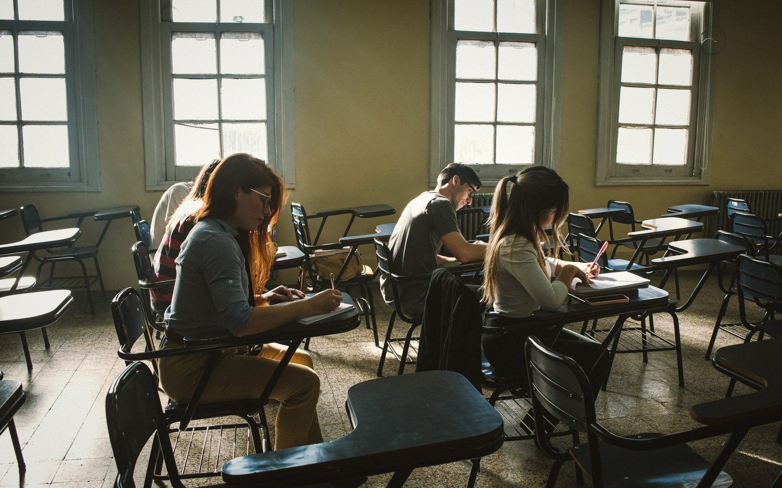 Students sit at scattered desks writing in a sunlit classroom